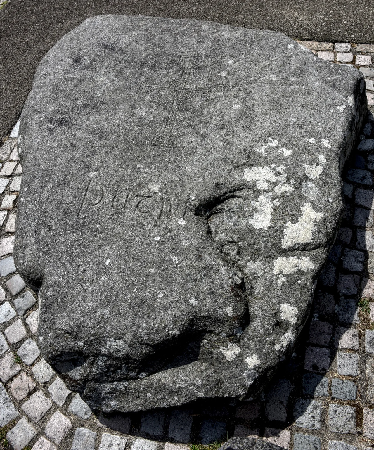 A rough granite stone bearing a carved cross and the name Patrick, surrounded by a cobbled border. St Patrick's traditional grave marker at Down Cathedral, photographed in July 2018.