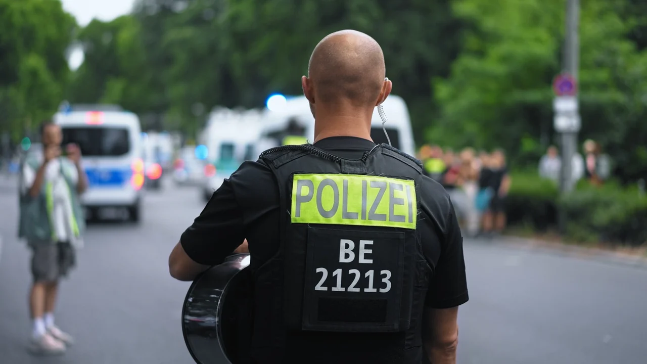 A German police officer seen from behind, wearing a vest marked POLIZEI, with patrol vehicles in the background.