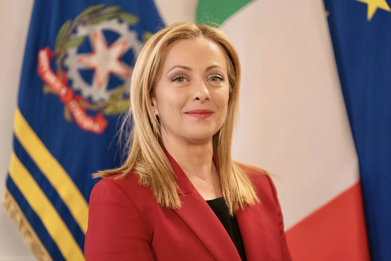 Giorgia Meloni in a red jacket in front of Italian and European flags, in an official portrait published in 2023.